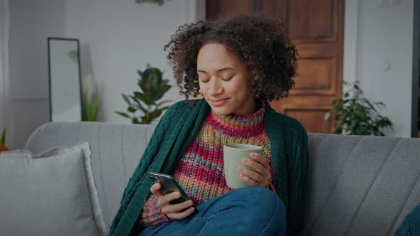 Young Peaceful Curly African American Lady Resting at Home Web Surfing on Smartphone and Enjoying