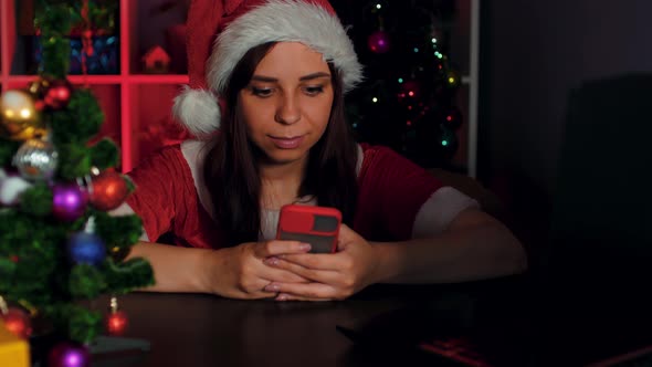 Young Woman in Santa Costume Using Smartphone in Office Sitting in Armchair in New Year's Atmosphere