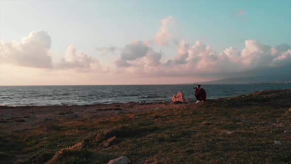 Photographer sitting on rocky beach and taking photos of pink beautiful sunset, wavy sea and sky