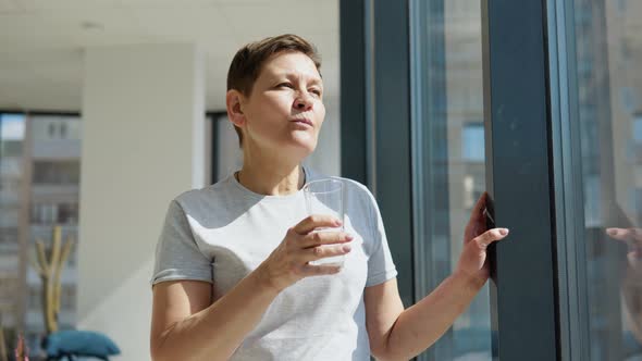 Senior Woman Drinks Water Standing Near the Window in a Light Room