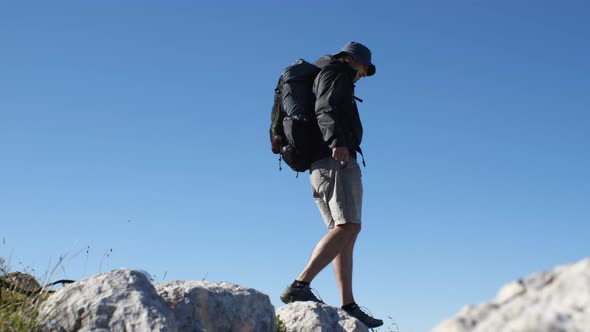 A man in shorts and a windbreaker with a backpack on his back jumps on rocks in the mountains agains