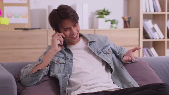 In his living room, an Asian man is on the phone, listening and conversing on his smartphone.