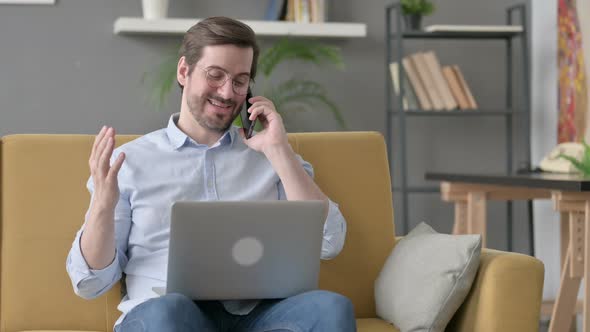 Young Man with Laptop Talking on Smartphone on Sofa