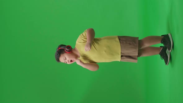 Side View Of Asian Little Boy Listening To Music With Headphones And Dancing On The Green Screen