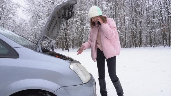 A Woman Calls on Her Smartphone While Standing By Broken Car with Open Car Hood in Winter
