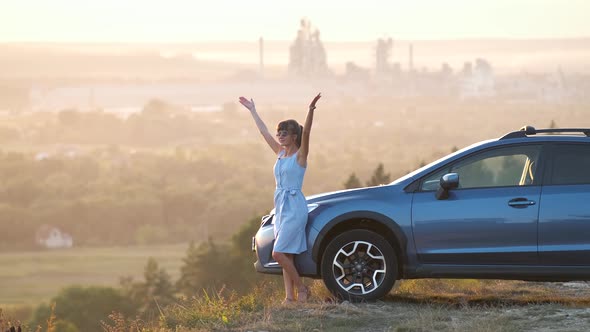 Happy young woman driver with outstretched up hands enjoying warm summer evening standing 