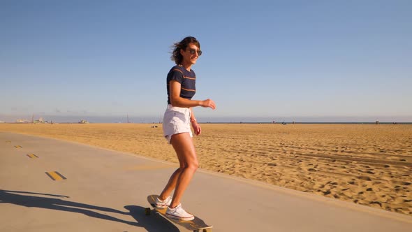 Sporty Young Woman In Sunglasses Dancing And Spinning While Riding Longboard In The Beach Park. - wi