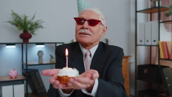 Senior Businessman Celebrating Lonely Birthday in Office Blowing Candle on Small Cake Making a Wish