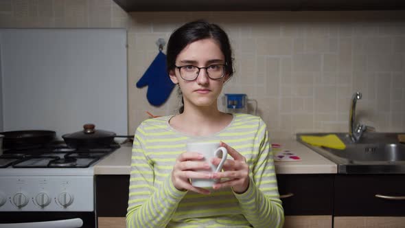 Girl with glasses is sitting at the kitchen table and drinking a hot drink from a white ceramic mug