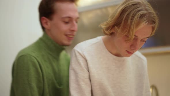 Blond guy cuts vegetables while his boyfriend fits hugging him