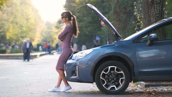 Puzzled female driver standing on a city street near her car with popped up hood looking 