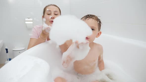 Cheerful Laughing Mother and Little Son Blowing Soap Bubbles and Foam While Playing in Bathroom