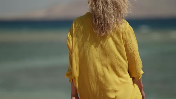 a Blonde in Sunglasses and a Yellow Fluttering Dress Walks Against the Background of a Blurred Sea
