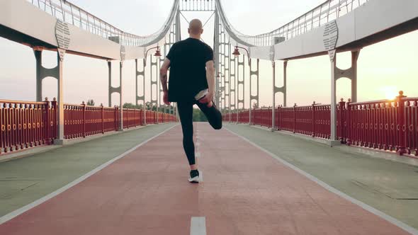 Man Runner in Black Sport Uniform Stretching Legs Before Sports on Pedestrian Bridge at Dawn