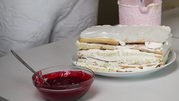 A Woman Smears A Biscuit Cake With Cream. Nearby Is A Container With Cherry Filling For The Cake.