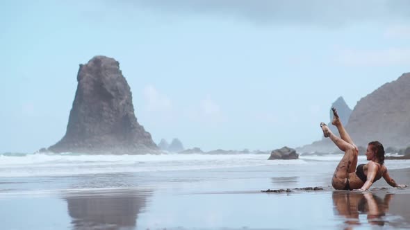 Woman Moves Plastically Dancing on the Sand Near the Ocean Against the Mountains and Black Sand