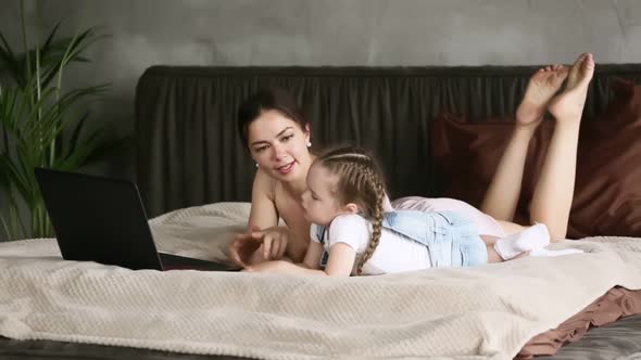 Mom Talking to Little Daughter Lying on Bed at Home with Laptop Iroi