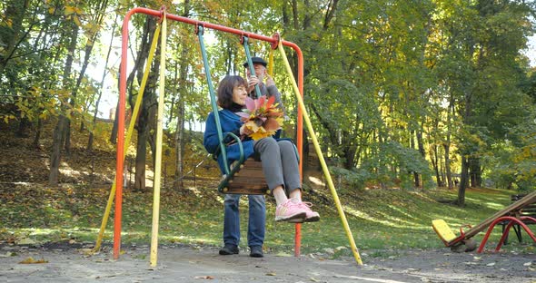 Bearded Husband Shakes a Rocking Chair with His Wife Holding a Bouquet of Autumn Leaves. Perky