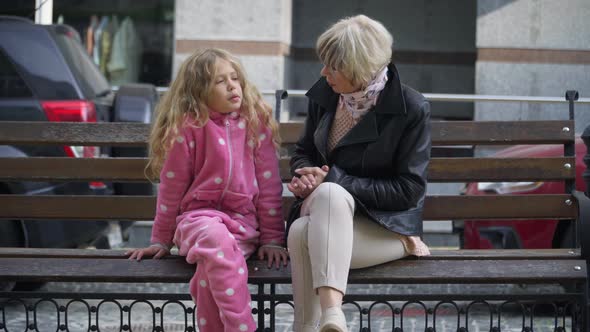 Cute Caucasian Girl in Pink Costume Sitting with Granny on City Street Bench Talking Smiling