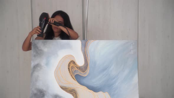 Female Artist Drying Top Side Of A Paint Canvas With A Hair Dryer In The Studio. medium shot