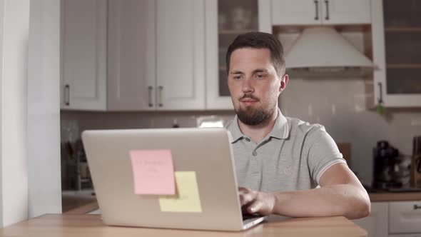 Adult Cheerful Man Is Working with Notebook in a Home, Sitting on a Kitchen