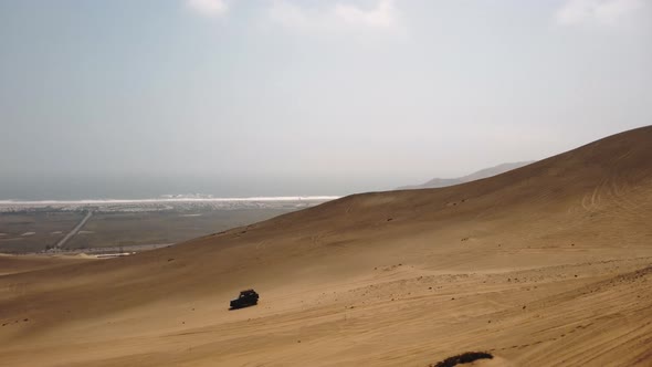 Aerial view of a 4x4 truck driving on sand dunes with sea background in Ica, Peru - tracking, drone