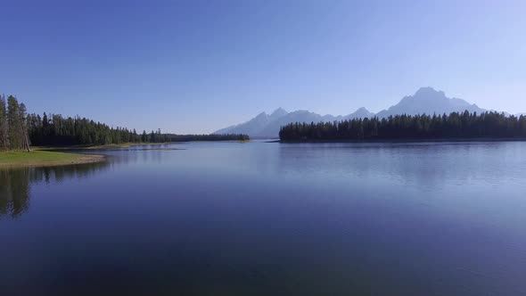 Camera flies over a calm Jackson Lake with mountains along the horizon AND PANS UP SLOWLY