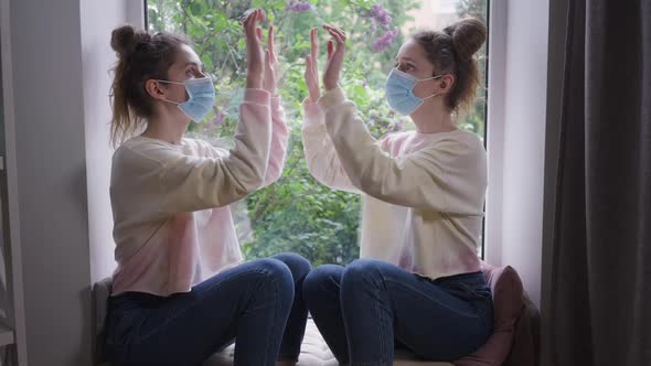 Identical Twins in Covid19 Face Masks Imitating Touching Glass Sitting on Windowsill at Home