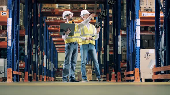 Two Workers with a Laptop Are Crossing the Warehouse Hall