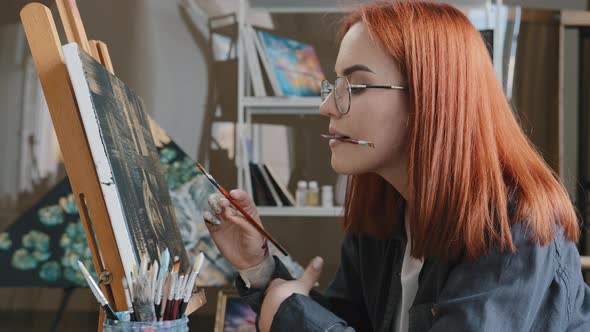 Inspired Funny Focused Artist Woman with Red Hair Girl Painter Wears Glasses Holds Painting Brushes