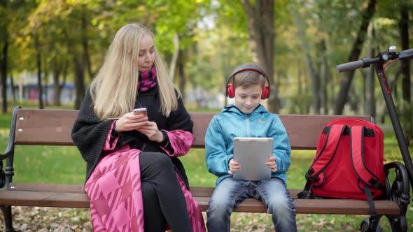 Absorbed Caucasian Young Mother and Little Son Surfing Internet on Smartphone and Tablet Sitting in