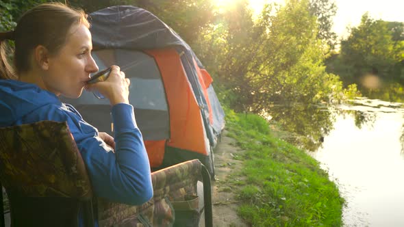 Woman is Drinking Tea in an Armchair Near a Tent on the River Bank