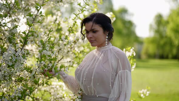 Attractive Brunette Woman in Vintage Dress is Walking in Blooming Garden in Spring Day