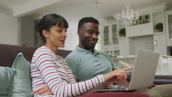 Happy diverse couple sitting on couch and using laptop in living room