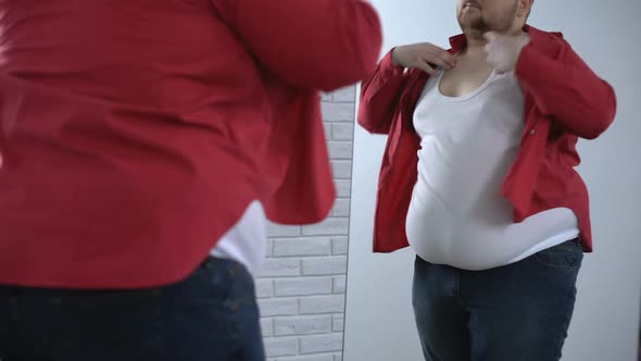 Overweight Male Putting Up Tight Red Shirt in Front Mirror, Preparing for Work
