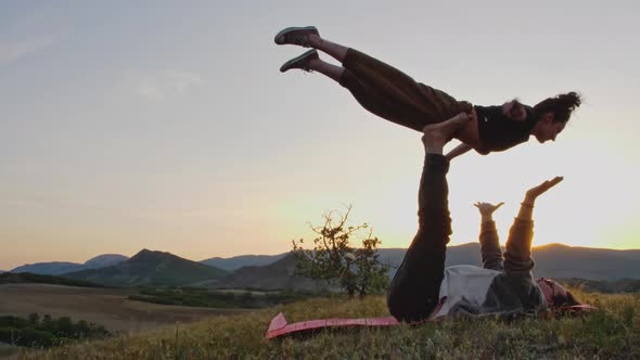 Young Man and Woman Do Aerial Yoga Exercise on Top of a Hill Against the Backdrop of Mountains