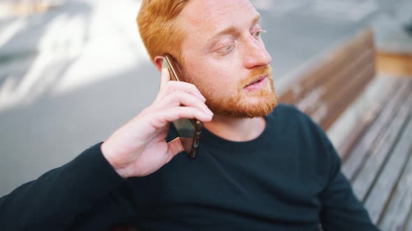 Handsome ginger man talking by smartphone while sitting on the bench