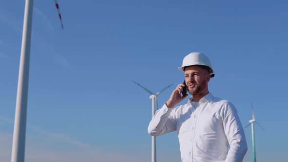 Engineer in Business Clothes and Helmet Talking on a Cell Phone Against the Backdrop of Wind