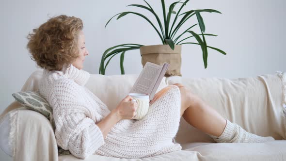 Woman Sitting On Sofa, Drinking Tea And Reading Book. Cozy Domestic Atmosphere