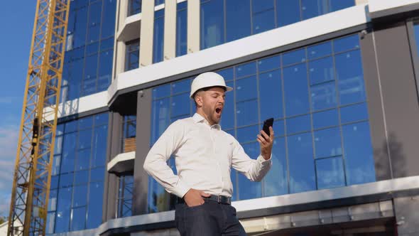 Engineer Architect in a White Shirt and Helmet on the Background of a Modern Glass Building Speaks