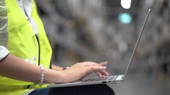 Asian female worker in safety vest sitting and working with computer laptop in storage warehouse. 