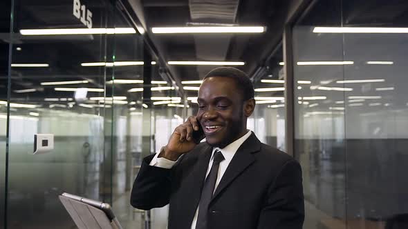 Professional Black-Skinned Office Manager Standing in Long Company Corridor and Talking on Phone