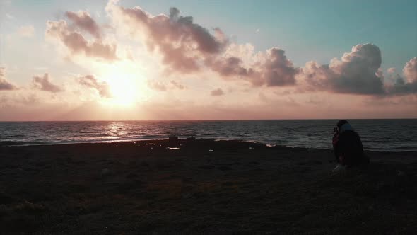 Female photographer on rocky seashore taking pictures and videos of pink sunset with sun reflection