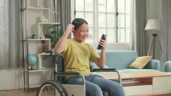 Happy Asian Kid Girl Sitting In A Wheelchair While Using Smart Phone And Celebrating At Home