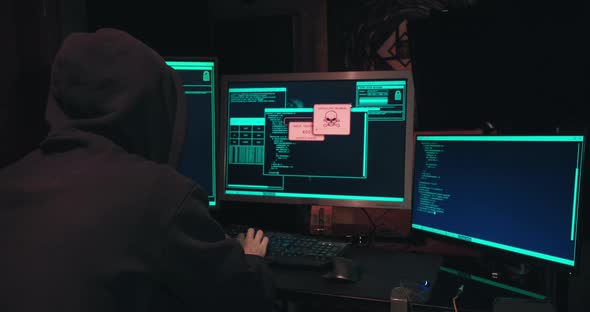 Hacker in a Hood Sits in Front of Computer Screens and Hacks Databases