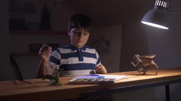 A Boy Paints While Sitting at a Table Under a Desk Lamp