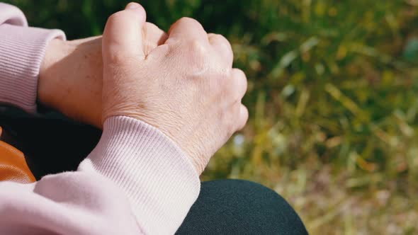 Elderly Woman Rubbing Wrinkled Hands Sitting in Nature in the Rays of Sunlight