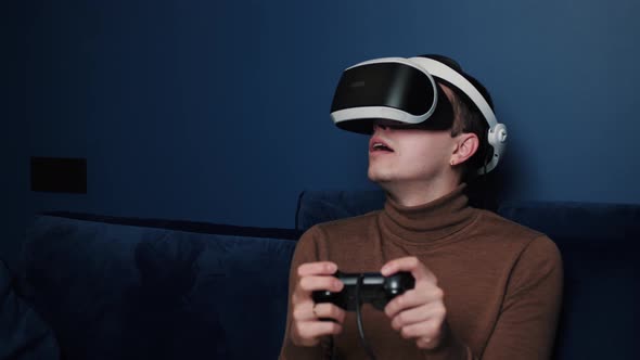 Attractive Caucasian Man Hold Joystick and Wear Virtual Reality Glasses Headset Helmet on Sofa at