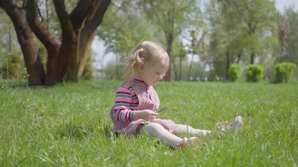 Cute Little Girl Sitting on the Grass in the Park, Playing Alone, Pointing with a Tiny Finger Up