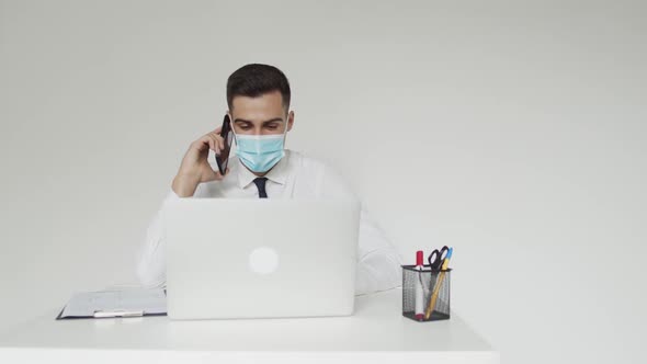 Office Manager in Medical Mask Talks on a Mobile Phone When Works on a Laptop